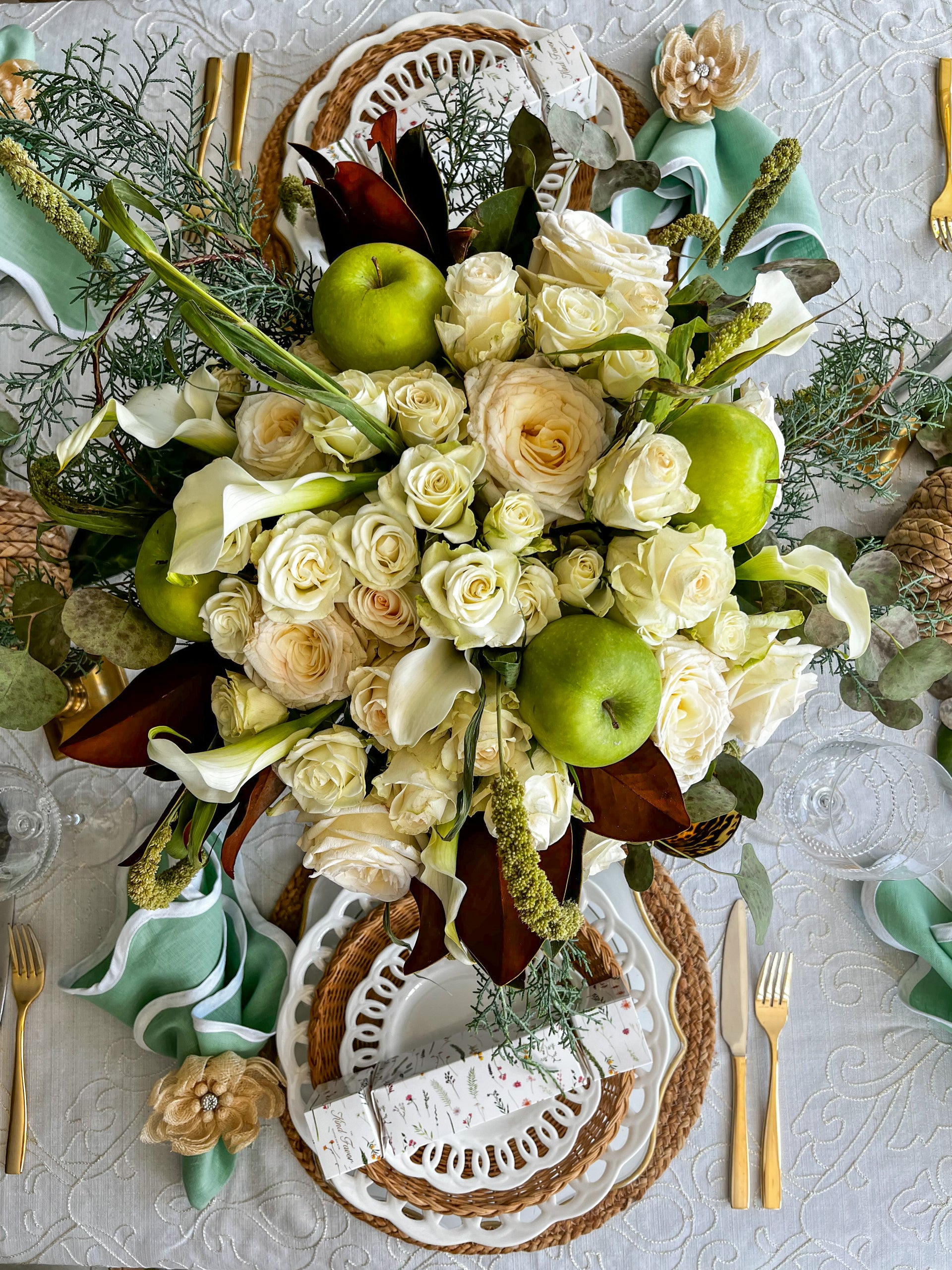 Overhead view of a lavish table centerpiece featuring white roses, green apples, and lush greenery, paired with a refined table setup including layered plates and gold utensils.