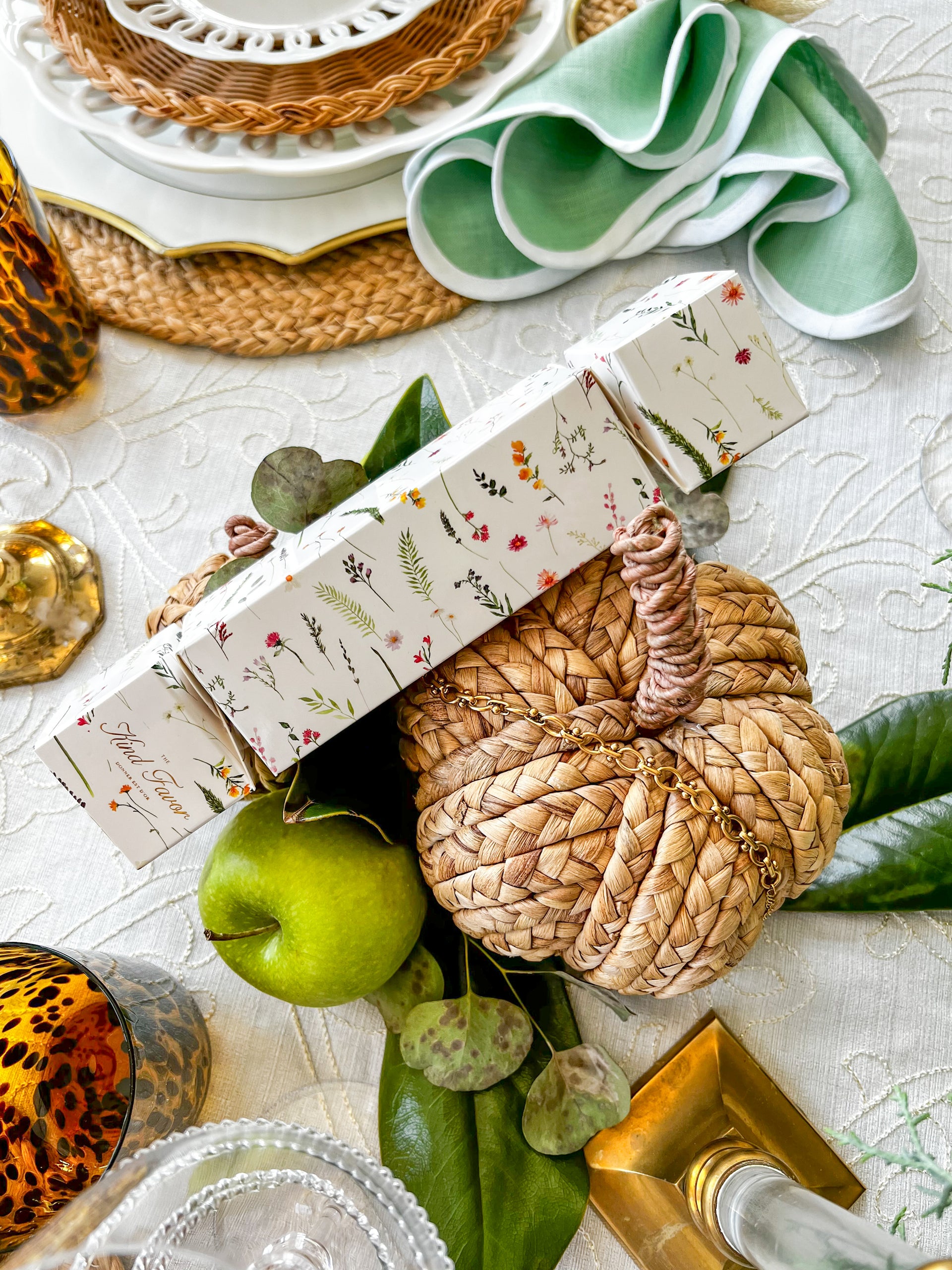 Festive holiday table decor with a woven pumpkin, green apples, and floral-patterned crackers, adding a rustic touch to the elegant setup.
