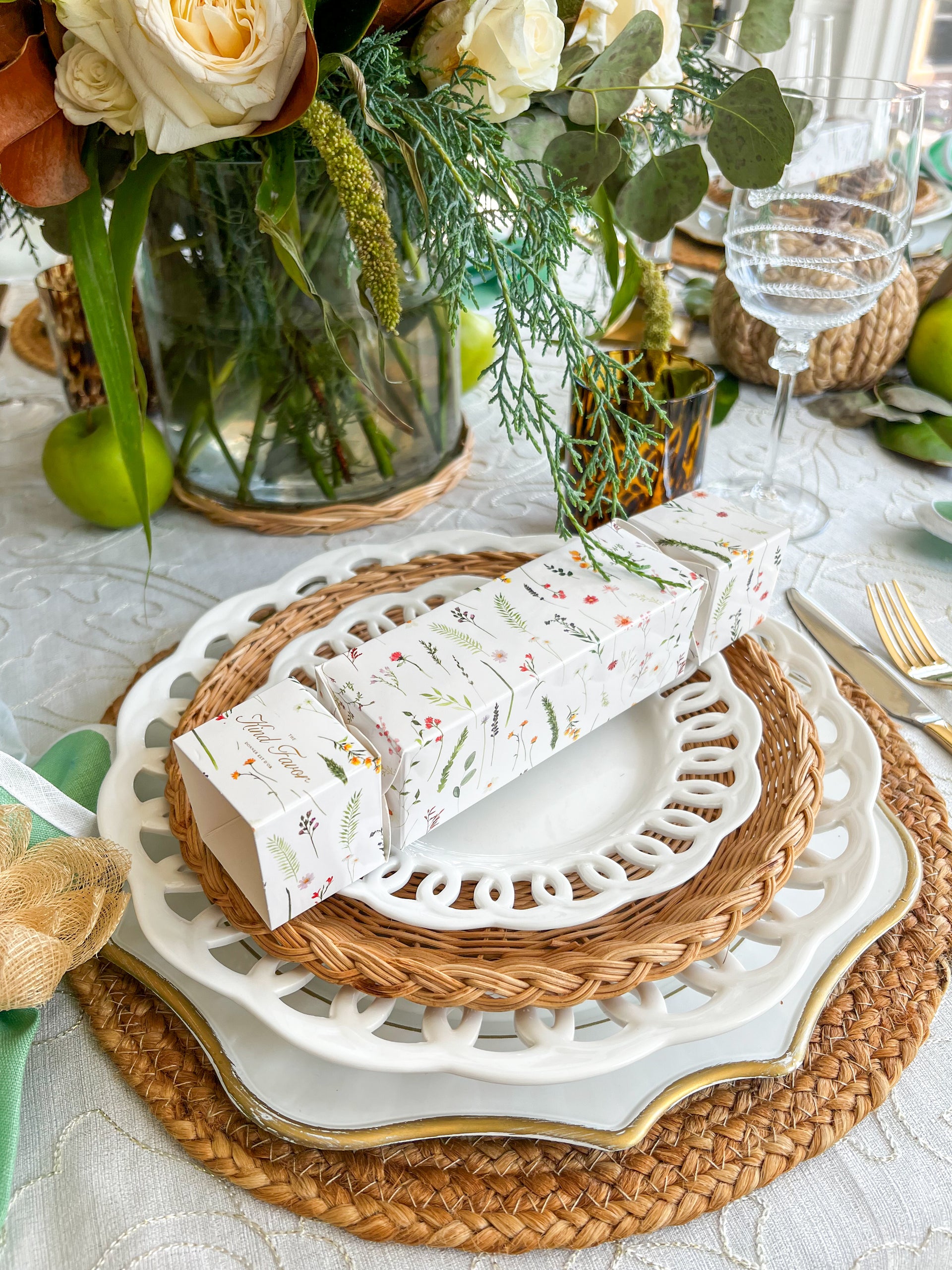 Close-up of a white floral-patterned party cracker on a layered plate setting with wicker and porcelain plates, highlighting the festive table details.